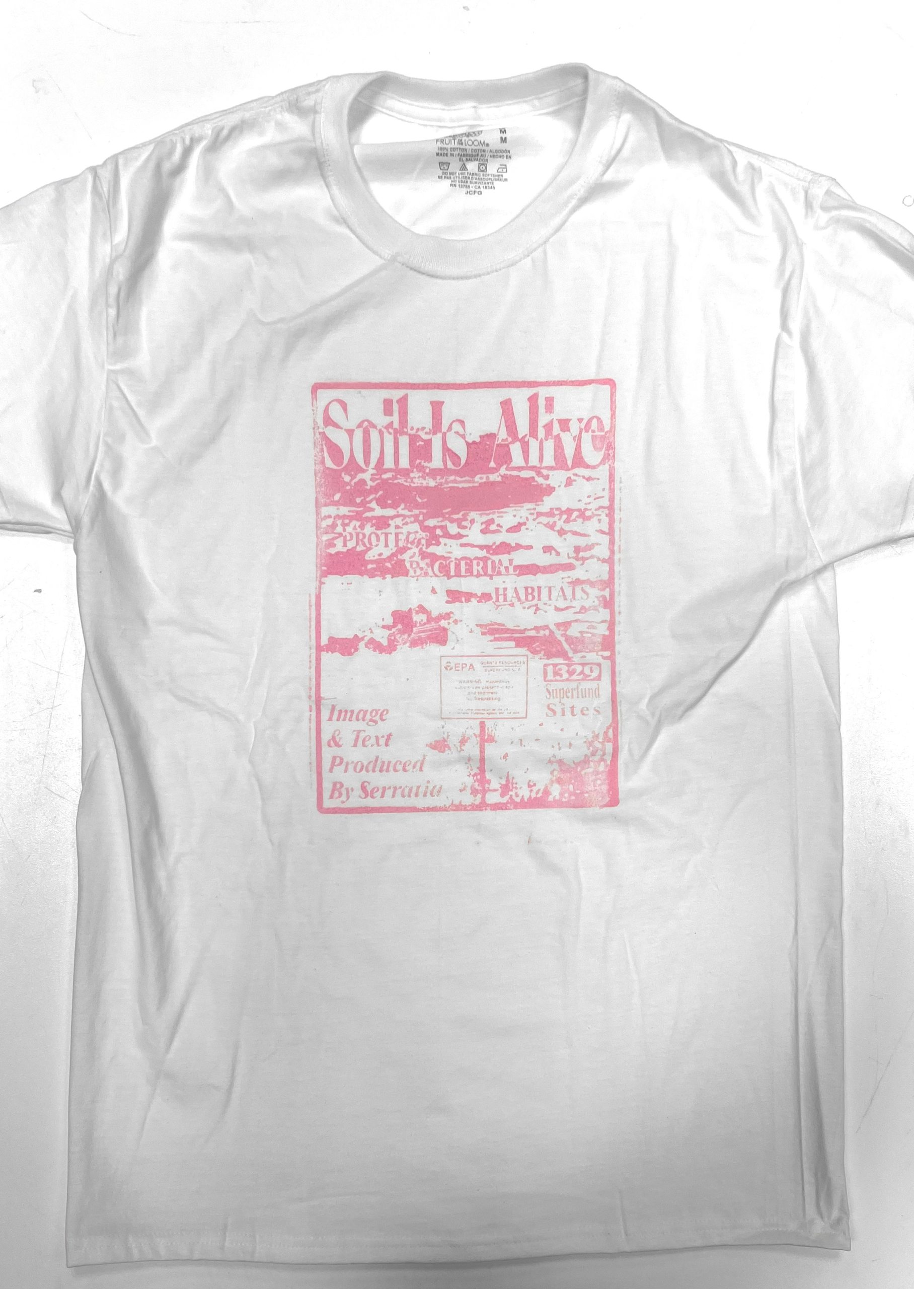 The finished white t-shirt laid flat, printed with the “Soil Is Alive” design in the pink pigment the cornstarch-thickened bacterial ink produced.
